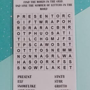 Può includere: Un cruciverba a tema natalizio su una carta bianca. La griglia contiene lettere per trovare parole come "regalo" e "pupazzo di neve". La carta è in una custodia trasparente, su uno sfondo azzurro chiaro.