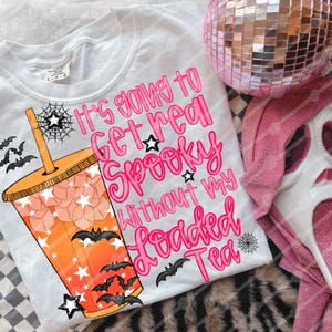 Puede incluir: Camiseta gris claro con el texto rosa "It's going to get real spooky without my loaded tea". La camiseta presenta un gráfico de una bebida naranja con estrellas y murciélagos. Una bola de discoteca rosa y una máscara rosa en el fondo.