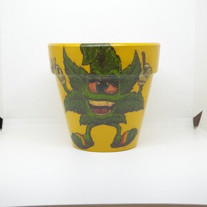 May include: Yellow ceramic flower pot featuring a cartoon illustration of a green marijuana leaf character wearing a hat and making a peace sign.
