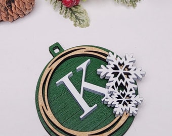 May include: A wooden Christmas ornament with a green background, a white letter "K", and a white snowflake. The ornament is round with a gold and brown border. Pinecones, red berries, and a garland with lights are in the background.