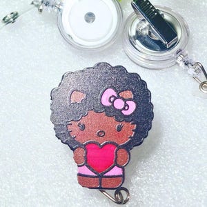 May include: A retractable badge reel featuring a cartoon character with dark skin, a pink bow, and a heart. The reel has a clear plastic casing and a metal clip. Green foliage is in the background.