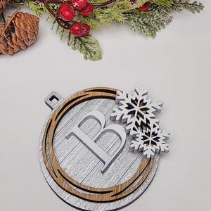 May include: Wooden Christmas ornament with a silver-painted background, a wooden border, and a white letter "B". Two white snowflake cutouts are attached. The ornament is a festive holiday decoration.