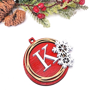 May include: A red wooden Christmas ornament with the letter "K" in white, surrounded by a light wood border. Two white snowflake cutouts are attached. Decorative pine and red berries are in the background.