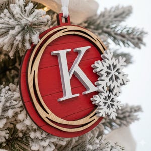 May include: Red and wood Christmas ornament with the letter "K" and snowflake accents. The ornament is round with a red center and a wooden border. It hangs from a white ribbon.