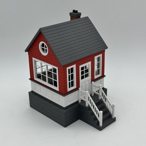 May include: A miniature model house featuring red horizontal siding, a dark gray shingled roof, and a dark gray brick-patterned foundation. The structure includes white window frames, a white door, white railings, and a set of stairs.