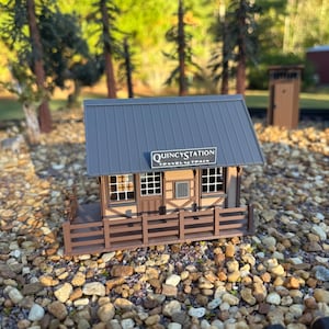 May include: A miniature model of a train station building with a dark gray metal-style roof, brown walls, and a front porch with a railing. A sign above the door reads Quincy Station Travel by Train in white lettering on a black background.