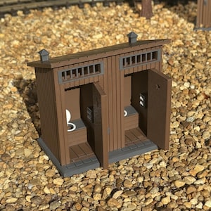 May include: A miniature brown double-stall outhouse model with vertical siding, two open doors, and gray trim. Each stall features a small white toilet seat, a toilet paper dispenser on the wall, and a rectangular vent window above the door.