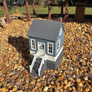 May include: A miniature model house featuring a blue exterior with white trim, a gray shingled roof, and a small chimney. The structure includes a front staircase with white railings, multiple windows, and a central door, all set on a gray foundation.