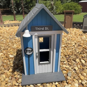 May include: A miniature blue and white building model with a gray gabled roof, featuring a sign that reads Third St. The structure includes a white door with a window, a small white light fixture, and a circular black and white stop sign.