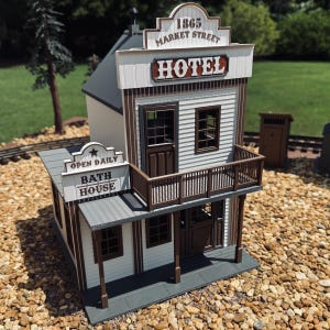 May include: A miniature model of a two-story building featuring white siding and brown trim. The upper level displays the text 1865 MARKET STREET HOTEL, while the lower section includes a sign reading OPEN DAILY BATH HOUSE.