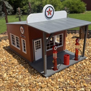 May include: A miniature Texaco gas station model featuring a brown brick building with a white roof sign, a gray canopy, and two red vintage-style gas pumps. The structure includes a white door, window frames, and a small red barrel on the porch.