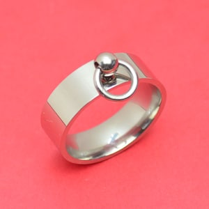 Könnte beinhalten: Ein silberfarbener Ring mit einem flachen Band und einem kleinen, kreisförmigen Ring, der an einer Kugel befestigt ist. Der Ring besteht aus einem glänzenden, metallischen Material und liegt auf rotem Hintergrund. Das Design des Rings ist schlicht und modern.