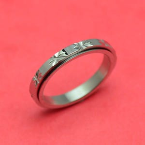 Könnte beinhalten: Ein silberfarbener Ring mit einem sich wiederholenden Sternmuster um das Band. Der Ring ist vor einem einfarbigen roten Hintergrund platziert. Der Ring hat ein schlichtes, elegantes Design.