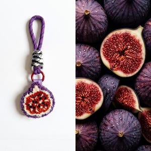 Pode incluir: Um porta-chaves em forma de figo com um design bordado em roxo e branco, um centro vermelho e sementes pretas. O pendente está preso a uma alça de cordão roxo com um nó estampado em preto e branco e um pequeno anel de metal vermelho.