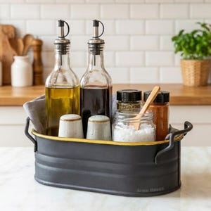 Pode incluir: Um carrinho de cozinha de metal preto com uma borda amarela contém duas garrafas de vidro de óleo e vinagre, potes de especiarias, sal e uma colher de pau. Dois pequenos saleiros também estão incluídos. O carrinho está em uma bancada branca.