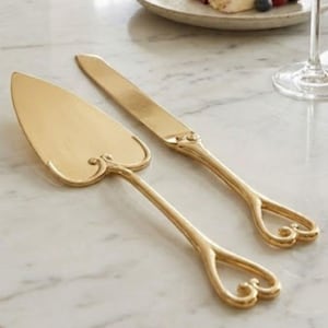 Könnte beinhalten: Ein goldfarbenes Tortenheber- und Messerset mit herzförmigen Griffdetails. Im Hintergrund liegt ein Stück Kuchen mit Beeren auf einem Teller, zusammen mit einem Glas Champagner. Das Set befindet sich auf einer Marmoroberfläche.