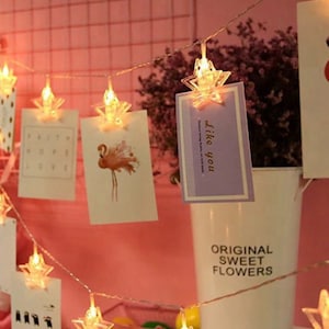 May include: A string of star-shaped LED lights with small clips holding up photos and cards. The cards have various images and text, including "Like you" and "Faith Hope Love". The background is a pink wall.