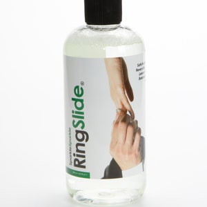 May include: A clear plastic bottle of RingSlide solution for removing stuck rings. The bottle has a black cap and a label with the RingSlide logo and the text "Sparkle Sparkle" in green. The label also features a photo of a couple's hands with a ring on the woman's finger.