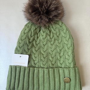 May include: A green cable-knit beanie with a brown faux fur pom-pom. The hat has a folded brim and a small gold-coloured emblem. A white tag is attached to the hat.