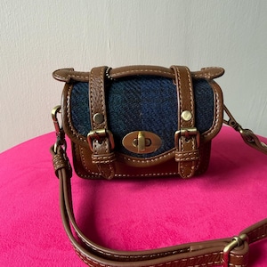 May include: A small satchel bag featuring a dark blue and black herringbone fabric centre panel with brown leather trim. The bag includes two decorative vertical leather straps with brass buckles, a central brass turn-lock closure, and a long shoulder strap.