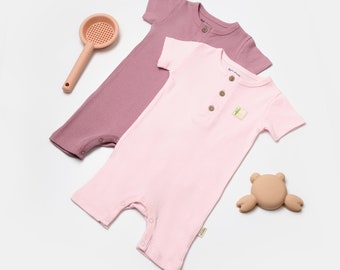 Puede incluir: Dos bodies de bebé, uno rosa y otro malva, con mangas cortas y cierres a presión. Los bodies están hechos de tela de canalé y tienen un pequeño parche en la parte delantera. También se ve un colador rosa y un juguete de goma marrón en la imagen.