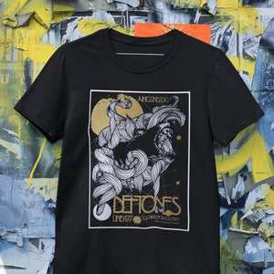 May include: Black t-shirt with a graphic print of a wolf's head with a stylized design. The text "Deftones" is printed in gold below the wolf's head, with the text "Line 477" and "Coachella 2010" printed in smaller gold text below the band name.