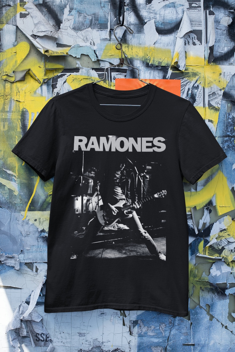 May include: Black t-shirt with a white graphic of a band playing on stage. The band's name "RAMONES" is printed in white above the image.