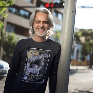 May include: A man with gray hair and a beard is wearing a black long-sleeved shirt with a graphic print of a black and white illustration of a creature with the text "MEMORIAS DE FIOS" in gold lettering.