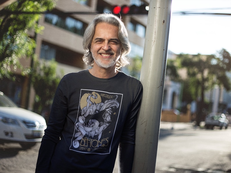 May include: A man with gray hair and a beard is wearing a black long-sleeved shirt with a graphic print of a black and white illustration of a creature with the text "MEMORIAS DE FIOS" in gold lettering.