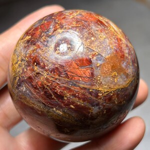 May include: A polished, spherical gemstone with a mix of reddish-brown and golden hues. The orb's surface displays intricate patterns and textures, creating a unique visual appeal. The gemstone is held in a hand, showcasing its size and shape.