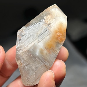 Könnte beinhalten: Ein klarer Quarz-Kristall mit inneren Streifen und orange-braunen Einschlüssen. Der Kristall wird in einer Hand gehalten, was seine natürliche, geometrische Form und seine durchscheinende Qualität zeigt. Die Facetten des Kristalls fangen das Licht ein und heben seine einzigartigen Muster hervor.