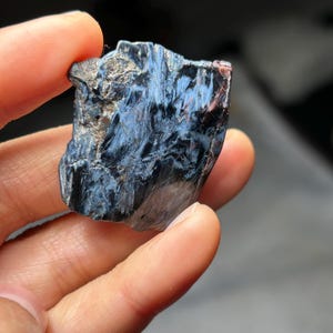 May include: A close-up of a piece of pietersite gemstone. The stone has a dark blue and black color with streaks of lighter blue and some reddish-brown hues. The gemstone is held in a hand.