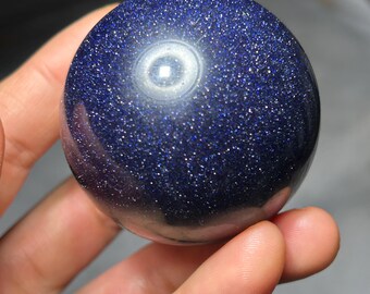 May include: A polished, spherical gemstone in a deep blue hue, speckled with shimmering inclusions. The sphere is held in a hand, showcasing its smooth, reflective surface. The gemstone's color and texture create a celestial appearance.