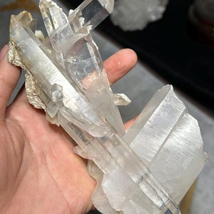 Könnte beinhalten: Ein Cluster aus klaren Quarzkristallen mit scharfen, spitzen Abschlüssen. Die Kristalle werden in einer Hand gehalten und zeigen ihre natürliche, durchscheinende Schönheit. Der Quarz sitzt auf einer beigen Matrix.
