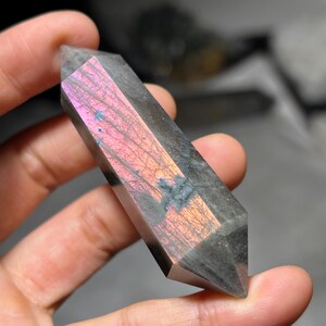 May include: A double-terminated labradorite crystal point featuring a dark gray base with a prominent, vibrant iridescent flash of pink, orange, and gold hues across its polished surface, showcasing natural internal fractures and mineral inclusions.