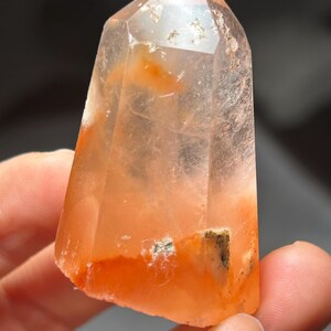 Könnte beinhalten: Ein klarer Quarz-Kristallpunkt mit orangefarbenen und rotbraunen Einschlüssen. Der Kristall wird in einer Hand gehalten und zeigt seine natürliche Form und Farbvariationen. Der Kristall hat eine polierte, facettierte Spitze und eine raue, natürliche Basis.