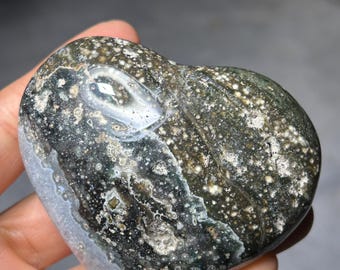 May include: A heart-shaped stone with a speckled pattern in shades of green, grey, and white. The polished surface has a smooth texture. The stone is held in a hand, showcasing its natural beauty and unique design.
