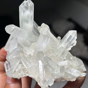 May include: A cluster of clear, translucent quartz crystals featuring multiple sharp, pointed terminations extending from a central base. The mineral specimen displays natural geometric facets and varying crystal sizes with a bright, glassy luster.