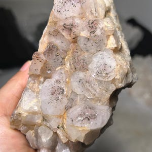Peut inclure: Un amas de cristaux de quartz translucides blanc rosé pâle, présentant de nombreuses petites inclusions minérales gris foncé à noires dispersées sur les surfaces facettées. Le spécimen minéral a une forme irrégulière et dentelée.