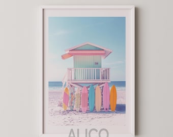 May include: A pastel-colored lifeguard stand on a sandy beach with a row of colorful surfboards leaning against it. The stand has a pink awning and a light blue and white striped door. The surfboards are pink, blue, yellow, and orange.