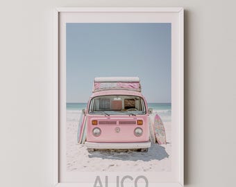 Op de afbeelding: Een roze vintage Volkswagen busje met surfplanken aan de zijkant geparkeerd op een zandstrand. De bus heeft een wit dak en een witte bumper. De lucht is blauw en de zee is op de achtergrond.
