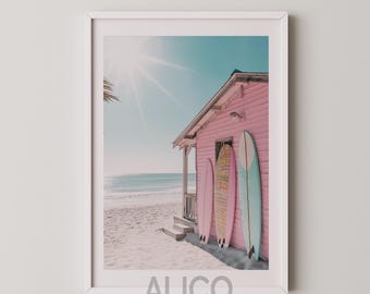 May include: A pink beach house with three surfboards leaning against the side. The surfboards are blue, pink, and yellow. The house is on a sandy beach with a blue ocean in the background.