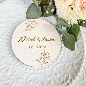 K&ouml;nnte beinhalten: Ein rundes Holzschild mit goldenem Blumendesign und dem Text "David & Laura 08.13.2023" auf wei&szlig;em Hintergrund.