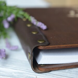 May include: A brown leather-bound journal with brass accents and a white paper interior. The journal is closed and lying on a white wooden surface.