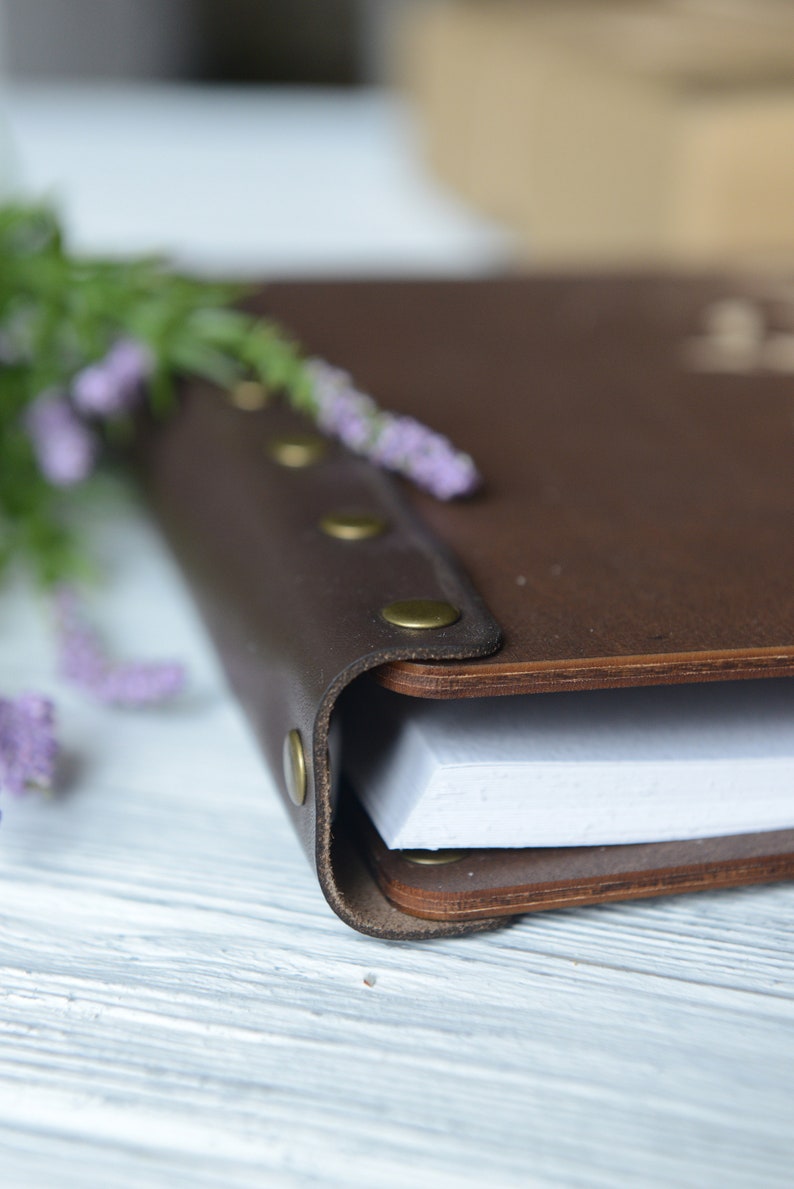 May include: A brown leather-bound journal with brass accents and a white paper interior. The journal is closed and lying on a white wooden surface.