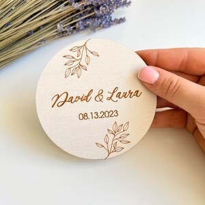 K&ouml;nnte beinhalten: Ein runder, h&ouml;lzerner Untersetzer mit hellbraunem Hintergrund und einem braunen, gravierten Design aus Bl&auml;ttern und dem Text "David & Laura 08.13.2023".