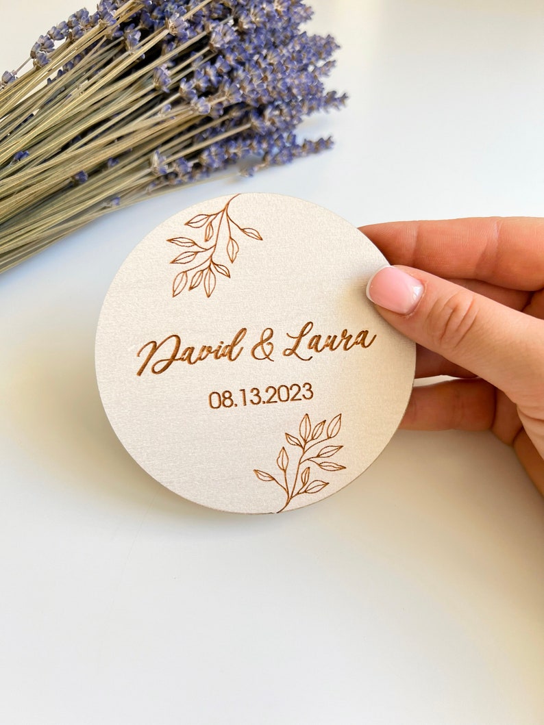 K&ouml;nnte beinhalten: Ein runder, h&ouml;lzerner Untersetzer mit hellbraunem Hintergrund und einem braunen, gravierten Design aus Bl&auml;ttern und dem Text "David & Laura 08.13.2023".