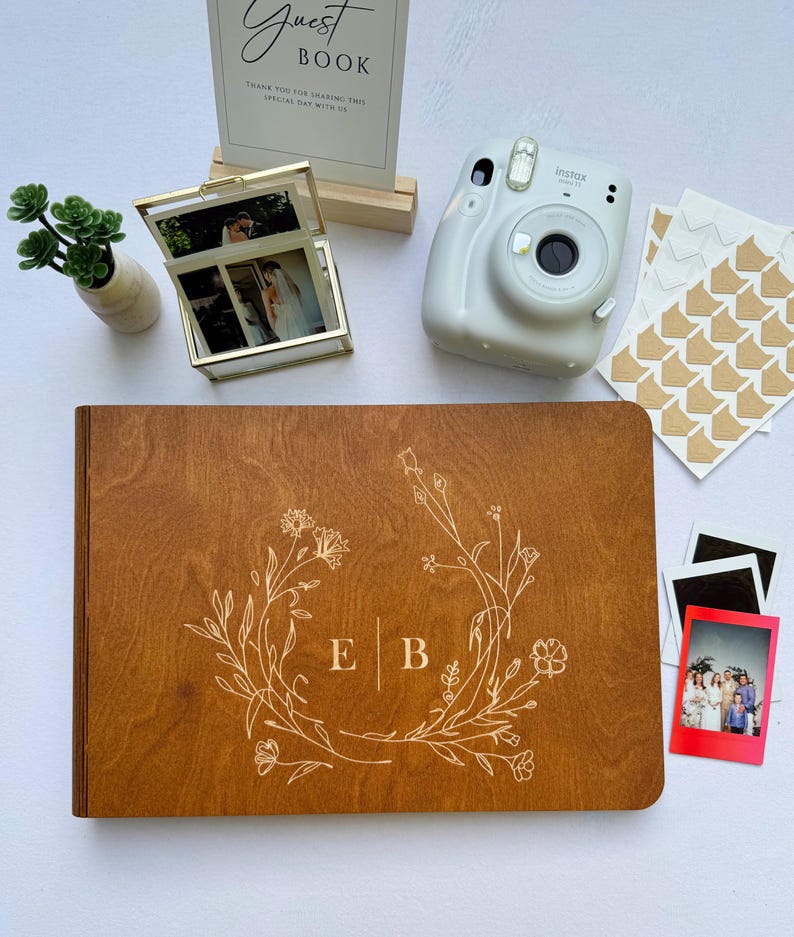 May include: A wooden guest book with floral design and initials, alongside a polaroid camera, photos, and a sign that reads "Guest Book". A small vase with greenery and a gold framed photo box are also present.