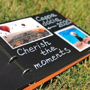 May include: A black scrapbook with a brown edge. The scrapbook has two photos inserted. The first photo shows a hot air balloon ride over a landscape. The second photo shows a beach scene. The scrapbook has the text "Cherish the moments" written on it. The text "Cappadocia 2020" is also written on the scrapbook.