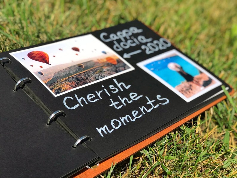 May include: A black scrapbook with a brown edge. The scrapbook has two photos inserted. The first photo shows a hot air balloon ride over a landscape. The second photo shows a beach scene. The scrapbook has the text "Cherish the moments" written on it. The text "Cappadocia 2020" is also written on the scrapbook.
